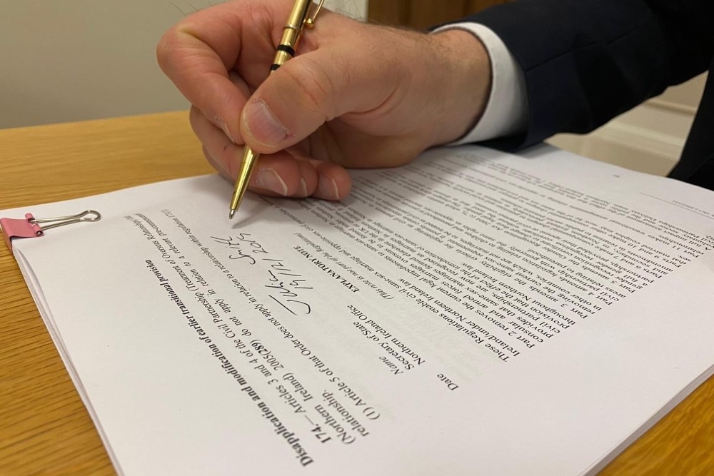 Image of a hand with a gold pen signing a document. The document being signed is the same-sex marriage regulations for Northern Ireland