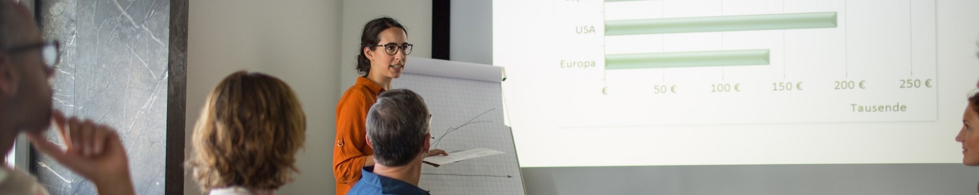 Person standing in front of a whiteboard and flip chart, presenting to an audience seated in a room, with gestures indicating key points from their presentation.